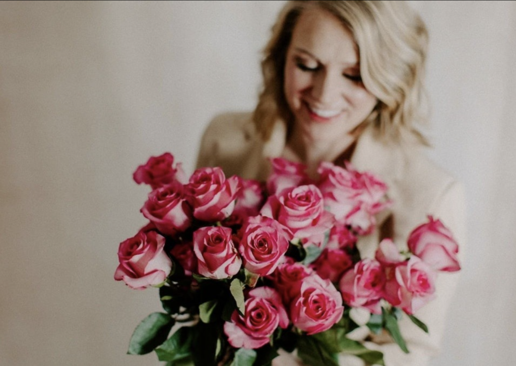 blonde girl in beige blazer smiling down holding pink roses 